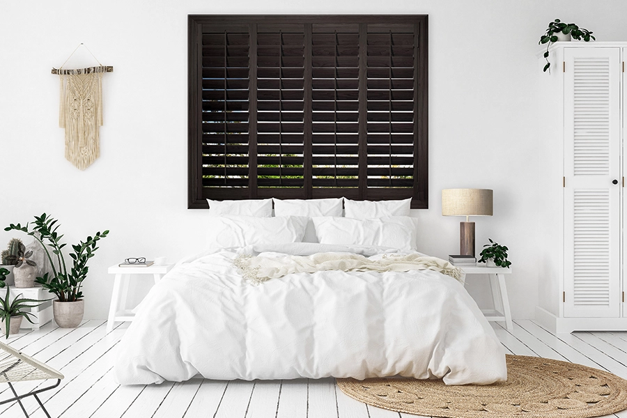 Quality wood shutters add class and a natural element to your home's decor. Dark wood Ovation shutters on a window in a white bedroom